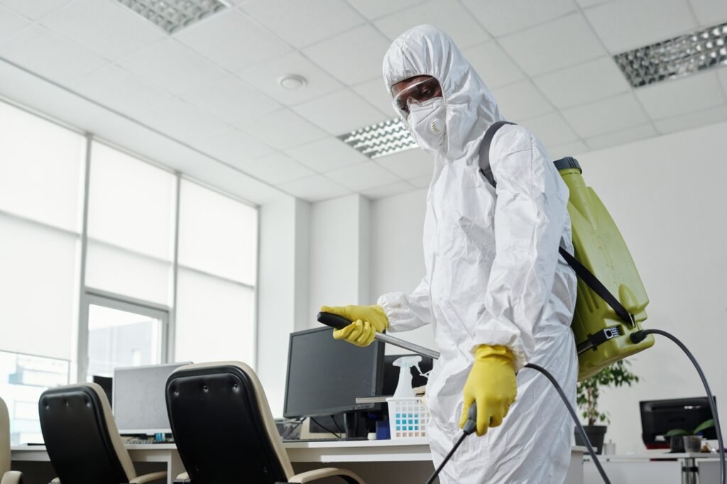 Black man in hazmat suit spraying floor of openspace office with disinfectant